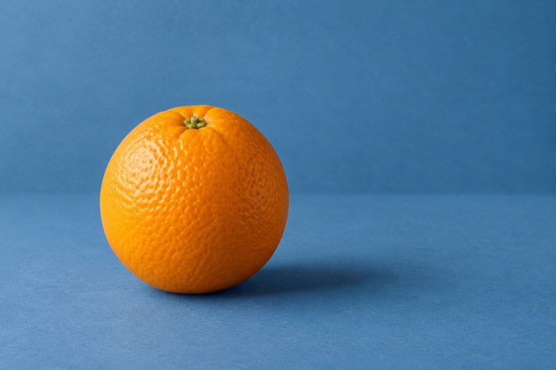 Uma única laranja sobre uma superfície azul suave diante de um fundo azul, com uma sombra macia.
